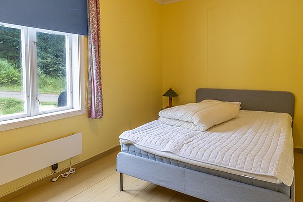 Gemütliches Schlafzimmer in Hitra, Norwegen, mit einem großen Bett und gelben Wänden. Ein Fenster bietet Blick ins Grüne, ideal für ruhige Nächte nach aufregenden Angelabenteuern. Perfekte Unterkunft für entspannte Angelreisen.