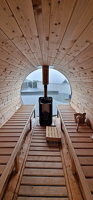 Gemütliche Fasssauna aus Holz in Südwest-Norwegen. Blick durch ein kreisrundes Fenster, Holzofen in der Mitte, Bänke auf beiden Seiten. Perfekt zum Entspannen nach einem erlebnisreichen Angelurlaub in Norwegen.
