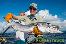 Ein Angler hält einen großen Barrakuda auf einem Boot vor maledivischer Meereskulisse. Der klare blaue Himmel und die leichte Brise versprechen ideale Bedingungen für einen erfolgreichen Angelurlaub. Perfekt für Abenteurer und Meeresangler.