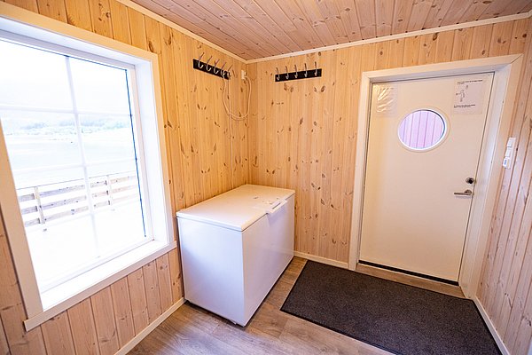 Gemütlicher Vorraum in einem norwegischen Angelurlaubshaus in Nordland mit Holzvertäfelung, Blick aufs Wasser durch großes Fenster, Tür mit Bullauge und Gefrierschrank für frisch gefangenen Fisch. Perfekt für erholsamen Angelurlaub.