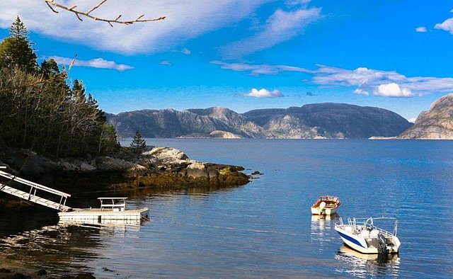 Küstenlandschaft in Hitra, Norwegen: Ruhiges Meer mit zwei Booten, umgeben von bewaldeten Felsen und majestätischen Bergen, ideal für Angelurlaub und Erholung in der Natur. Klarer Himmel und sanfte Wellen versprechen entspanntes Meeresangeln.