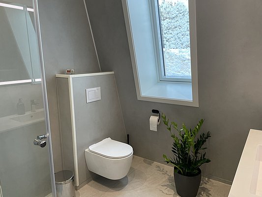 Modernes Badezimmer in Hitra, Norwegen, mit wandhängendem WC, großen Fenster für natürliches Licht, grauen Wänden und grüner Pflanze. Idealer Ausgangspunkt für Angelabenteuer und entspannte Erholung in skandinavischem Design.
