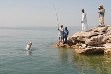Mehrere Personen stehen auf einem felsigen Ufer am Wasser, während eine Person einen großen Fisch angelt, der aus dem Wasser springt.