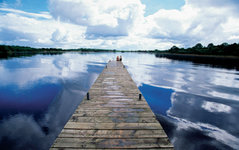Eine Holzbrücke führt über einen ruhigen, wolkengesäumten See, unter einem weiten blauen Himmel.
