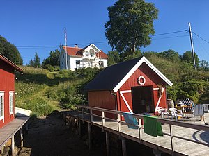 Rotes Bootshaus und weißes Haus auf grünem Hügel bei strahlend blauem Himmel in Hitra, Norwegen. Ideal für Angelreisen und Erlebnisurlaub in idyllischer Natur. Perfekte Mischung aus Komfort und Abenteuerlust.