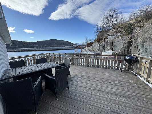 Eine sonnige Holzterrasse mit Essbereich und Grill bietet entspannte Aussicht auf Fjord und Berge in Hitra, Norwegen. Perfekt für einen Angelurlaub in idyllischer Umgebung.