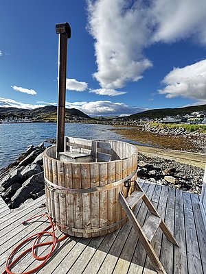 Holzfass mit Schornstein auf hölzerner Terrasse an norwegischem Fjord. Malerische Landschaft mit Bergen und Himmel. Perfekt für entspannte Angelreisen in Nord-Norwegen. Ideal für Naturliebhaber und Abenteuerlustige.