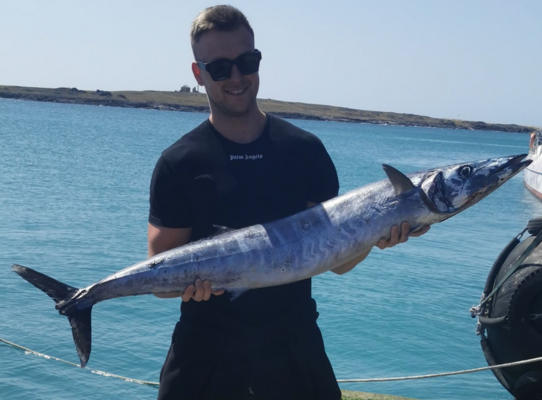 Ein Angler in Sao-Vicente, Kapverden, hält stolz einen großen Wahoo vor der malerischen Küstenlandschaft, ideal für Angelreisen und Meeresabenteuer.