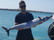 Ein Angler in Sao-Vicente, Kapverden, hält stolz einen großen Wahoo vor der malerischen Küstenlandschaft, ideal für Angelreisen und Meeresabenteuer.