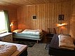 Gemütliches Holzinterieur in nordnorwegischer Hütte mit drei Einzelbetten, warmem Licht und grünen Vorhängen. Idealer Rückzugsort für Angelreisen in Norwegen, bietet Komfort und Entspannung nach einem erfolgreichen Angeltag.