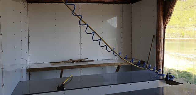 Offene, lichtdurchflutete Werkstatt in Nordland, Norwegen. Moderne Ausstattung mit Luftschlauch, helle Wände, Holzbalken, große Fenster mit Blick auf die grüne Landschaft. Ideal für Angelabenteuer und handwerkliche Projekte umgeben von Natur.