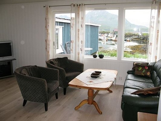 Gemütliches Wohnzimmer mit Blick auf einen norwegischen Fjord in Nordland. Helle Sitzecke, moderne Möbel, große Fenster und wunderschöner Ausblick auf Boote und Berge. Perfektes Angelreiseziel für Erholung und Abenteuer.