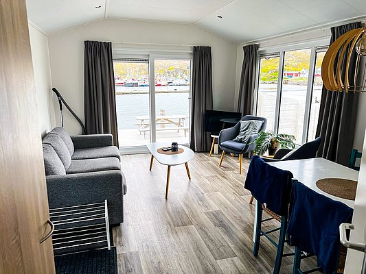 Gemütliches Wohnzimmer mit grauem Sofa und Sesseln in Nord-Norwegen. Große Fenster bieten Blick auf einen malerischen Hafen. Helle, moderne Einrichtung schafft eine einladende Atmosphäre für Angelreisende. Perfekter Rückzugsort nach einem erfolgreichen Angeltag.
