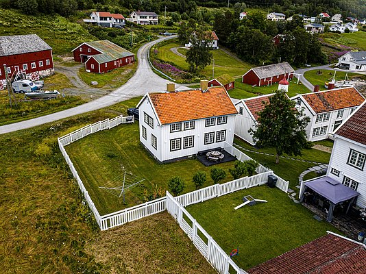 Charmantes Dorf in Nord-Norwegen: Traditionelle weiße Holzhäuser mit roten Dächern, umgeben von grünen Wiesen und Gärten. Malerische Landschaft mit blühenden Bäumen und ruhigen Straßen. Ideal für Angelurlauber und Naturliebhaber.
