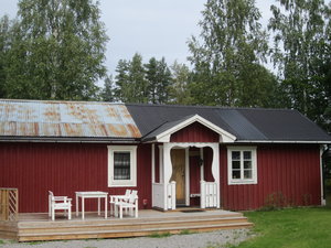 Rotes Holzhaus mit rustikalem Blechdach in Hälsingland, Schweden. Umgeben von grünen Bäumen, lädt die Veranda mit weißen Möbeln zum Verweilen ein. Perfekt für entspannte Angelurlaube in idyllischer Natur.