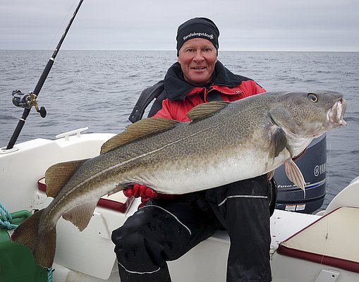 Ein Angler hält einen beeindruckenden Kabeljau auf einem Boot vor der Küste Nord-Norwegens. Im Hintergrund erstreckt sich das weite Meer unter einem bewölkten Himmel. Perfekter Ort für Angelabenteuer in Norwegen.