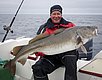 Ein Angler hält einen beeindruckenden Kabeljau auf einem Boot vor der Küste Nord-Norwegens. Im Hintergrund erstreckt sich das weite Meer unter einem bewölkten Himmel. Perfekter Ort für Angelabenteuer in Norwegen.