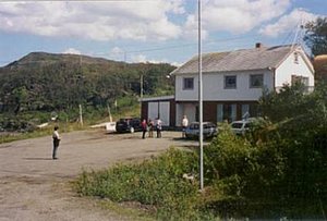Ländliche Szene in Nord-Norwegen: Ein Parkplatz vor einem zweistöckigen Gebäude, umgeben von grüner Landschaft und bewölktem Himmel, bietet einen authentischen Einblick in die ruhigere Seite Norwegens. Perfekt für Angelurlaube.