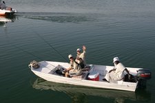 Drei Personen sitzen in einem kleinen Motorboot auf einem ruhigen Gewässer und halten Angelausrüstung, während sie fröhlich winken.