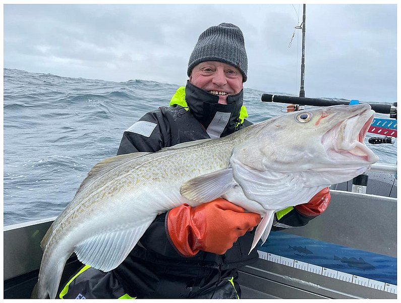 Ein Angler hält auf einem Boot einen großen Kabeljau vor einer stürmischen Meereskulisse.