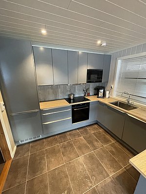 Moderne, gut ausgestattete Küche in einem Ferienhaus in Südwest-Norwegen. Graue Schränke, Holzarbeitsplatten und Fliesenboden bieten Komfort und Funktionalität. Ideal für Angelurlauber, die nach einem erfolgreichen Tag entspannen möchten.