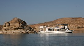 Ein weißes Boot fährt auf einem ruhigen See, flankiert von einer felsigen Insel und einer kargen, hügeligen Landschaft unter einem strahlend blauen Himmel.
