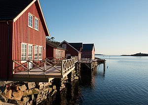 Rote Fischerhütten am ruhigen Wasser in Hitra, Norwegen, unter blauem Himmel. Idealer Ausgangspunkt für Angelreisen und Naturgenuss. Malerische Küstenlandschaft lädt zum Entdecken ein. Perfekt für Erholung und Abenteuer.