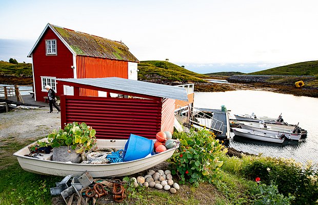 Küstenidylle in Nordland, Norwegen: Rotes Fischerhaus, Boote am Steg und Natur laden zum Angelurlaub ein. Grüne Hügel und das Wasser bieten eine malerische Kulisse für Abenteuer und Entspannung.