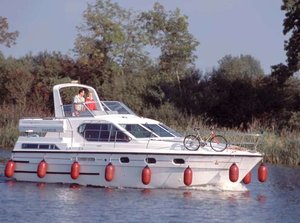 Ein weißes Hausboot fährt gemächlich auf dem River Shannon in Irland entlang, umgeben von üppiger Natur. Ein Fahrrad ist an Bord, was Abwechslung von der Wasserreise verspricht. Idyllische Angelreise.