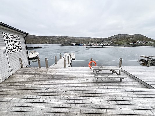 Holzdeck am Skarsvåg Turistcenter in Nord-Norwegen, mit Blick auf ruhige Hafenlandschaft, Boote und umliegende Hügel. Perfekter Ausgangspunkt für Angelreisen und Erkundungen in der arktischen Landschaft.