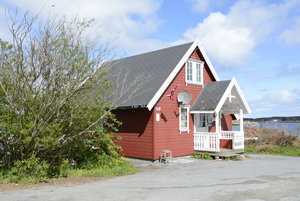 Rotes Ferienhaus in Hitra, Norwegen. Traditionelles skandinavisches Design, umgeben von grünen Bäumen. Nähe zum Meer, ideal für Angelreisen und Erholung. Blauer Himmel und ruhige Landschaft bieten Entspannung und Abenteuer.