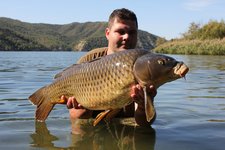 Ein Angler steht knietief im Wasser eines Sees und hält stolz einen großen Karpfen in seinen Händen, während im Hintergrund bewaldete Hügel zu sehen sind.