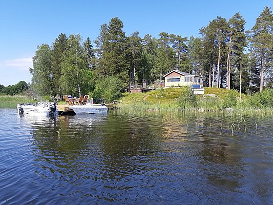 Ein idyllisches, mit Bäumen umgebenes Haus an einem Seeufer mit zwei Booten an einem kleinen Steg im Vordergrund.
