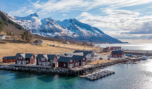 Malerische Landschaft in Nordland, Norwegen: Rote Fischerhütten am klaren Wasser, umgeben von schneebedeckten Bergen und blauem Himmel. Perfekt für Angelurlaub in einer beeindruckenden Naturkulisse.