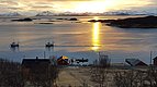 Nord-Norwegens Küste bei Sonnenuntergang: Fjordlandschaft, schneebedeckte Berge, ruhige See mit Fischerbooten, traditionelle rote Holzhäuser im Vordergrund. Beliebtes Ziel für Angelurlaub und Abenteuerreisen. Ideal zum Meeresangeln und Erleben unberührter Natur.