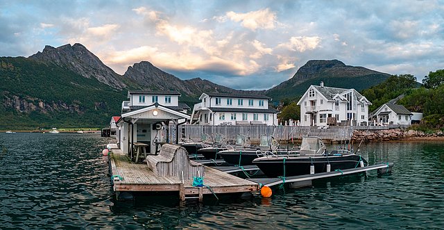 Norwegisches Nordland: Malerischer Fjordhafen mit Anlegesteg, umgeben von majestätischen Bergen und charmanten weißen Häusern. Ideal für Angelurlauber, die Ruhe und atemberaubende Landschaften suchen. Perfekter Ausgangspunkt für Angeltouren und Naturentdeckungen.