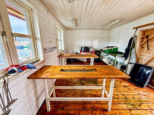 Helle Fischverarbeitungsstation in Nord-Norwegen mit rustikalen Holztischen, weißer Holzverkleidung und Hafenblick. Perfekt für Angler, um den frischen Fang effizient zu säubern und vorzubereiten. Ideal für Angelurlauber in Norwegen.