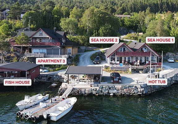 Luftaufnahme einer idyllischen Küstenunterkunft in Südwest-Norwegen mit traditionellen roten Holzhäusern, eigenem Bootssteg und Hot Tub. Perfekt für Angelurlauber, die Erholung und Abenteuer inmitten beeindruckender Natur suchen. Keywords: Südwest-Norwegen, Angelreise, Küstenunterkunft.