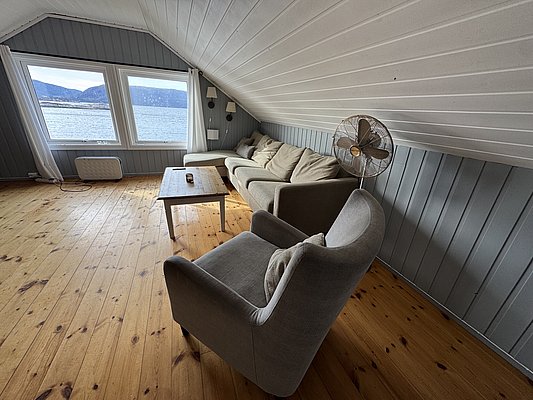 Gemütlicher Wohnbereich mit grauem Sofa, Holzfußboden und großen Fenstern mit Meerblick in Hitra, Norwegen. Ideal für entspannenden Angelurlaub. Heizung und Ventilator sorgen für Komfort bei jedem Wetter.