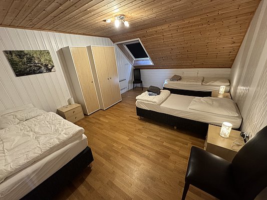 Gemütliches Schlafzimmer in Hitra, Norwegen mit Holzdielen, drei Betten, Dachschräge und sanfter Beleuchtung. Ideal für erholsame Angelurlaube mit Freunden. Entspannte Unterkunft nach erfolgreichen Tagen beim Meeresangeln.
