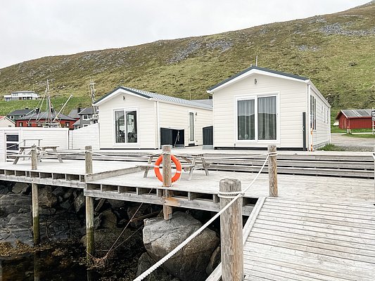 Holzterrasse mit zwei weißen Hütten in Nord-Norwegen, idyllisch am Wasser gelegen. Perfekte Ausgangsbasis für Angelreisen. Im Hintergrund grüne Hügel, ideal für Erholung und Abenteuer.