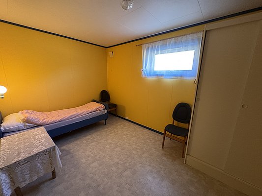 Das Bild zeigt ein einfaches, gemütliches Schlafzimmer mit gelben Wänden, einem Einzelbett, Tisch mit Spitzentischdecke und Stuhl in Hitra, Norwegen. Perfekt für entspannte Angelreisen.