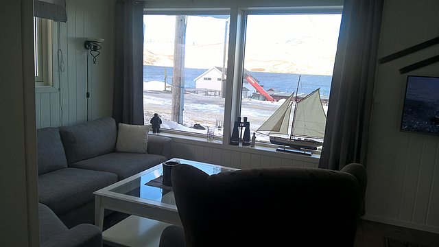 Gemütliches Wohnzimmer in Nord-Norwegen mit Blick auf schneebedeckte Landschaft und Meer. Maritime Dekorationen und helle Einrichtung sorgen für eine einladende Atmosphäre. Perfekter Ausgangspunkt für Angelreisen und Outdoor-Abenteuer in arktischer Natur.