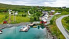 Luftaufnahme von Nord-Norwegen: malerisches Dorf mit bunten Häusern, Hafen und Booten an ruhiger Küste, umgeben von grüner Landschaft. Perfekt für Angelreisen und Naturerlebnisse.