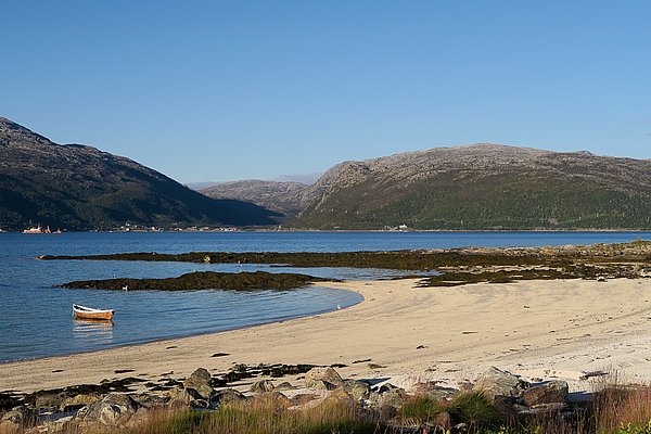 Sandstrand in Nord-Norwegen bei klarem Himmel, umrahmt von Bergen und ruhigem Wasser. Ein einsames Boot am Ufer verstärkt die idyllische, friedliche Atmosphäre. Perfekter Ort für einen entspannten Angelurlaub.