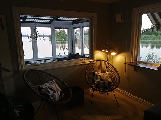 Ein gemütlicher Innenbereich mit zwei runden Korbstühlen und geometrischen Kissen wird von sanftem Lampenlicht erleuchtet, während der Blick durch ein großes Fenster eine ruhige Seeszene mit Bäumen im Abendlicht zeigt.