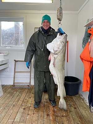 Ein Angler in Nord-Norwegen hält einen beeindruckenden Kabeljau in einer Hütte. Perfekter Fang im Angelurlaub, passende wetterfeste Kleidung, draußen verschneite Landschaft sichtbar. Abenteuerliches Meeresangeln in skandinavischer Umgebung.