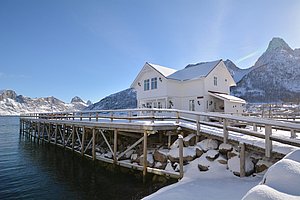 Schneebedecktes Holzhaus in Nord-Norwegen am klaren Fjord, umgeben von majestätischen, schneebedeckten Bergen unter strahlend blauem Himmel. Perfekte Kulisse für Angelabenteuer und naturverbundene Erholung.