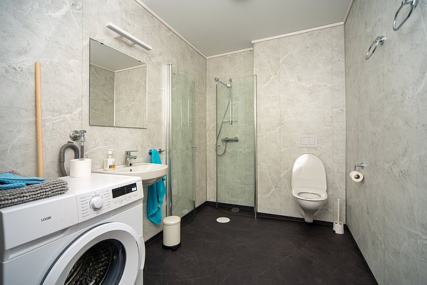 Modernes Badezimmer in Hitra, Norwegen, mit Waschmaschine, Dusche und WC. Helle Fliesen sorgen für ein einladendes Ambiente. Ideal für Angelurlauber, um Komfort und Funktionalität nach einem erfolgreichen Tag am Wasser zu genießen.