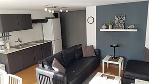 Moderne Ferienwohnung in Riba Roja, Spanien: Offener Wohnbereich mit schwarzem Sofa, Küchenzeile, dekorativem Regal und Esstisch. Ideal für Angelurlaub und Entspannung. Helle Atmosphäre für einladende Aufenthalte in der Natur.